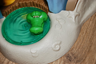 A green plastic toy hippopotamus with googly eyes floats in a small, round, green paddling pool on a moulded white plastic Tiki Hideout playset inspired by Jake and the Never Land Pirates, all placed on a wooden surface.