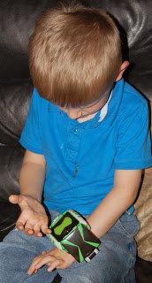 A young boy with blonde hair, wearing a blue shirt and jeans, sits on a dark sofa while looking down at a green and black toy watch on his wrist, clearly fascinated by one of his favourite toys.