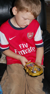 A young boy wearing a red Arsenal football shirt sits and looks down, thoughtfully holding one of his favourite toys—a yellow toy car—in his hands.