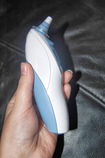 A hand holding a Braun ThermoScan ear thermometer with a blue and white casing, positioned against a dark leather background.