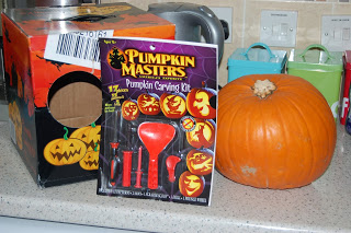 A Pumpkin Carving Kit from Waitrose, featuring various tools and sample designs, is displayed next to a large pumpkin and a decorated box with jack-o’-lantern faces on a kitchen worktop.