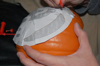 A person holds a pumpkin with a paper stencil taped to it, using a tool from a Pumpkin Carving Kit to outline a design for carving—perfect for your next Waitrose-inspired project or review.