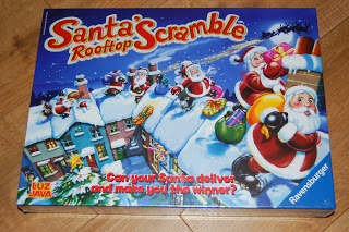 Santa's Rooftop Scramble board game box shows cartoon Santas delivering gifts on snowy rooftops at night, with text asking, "Can your Santa deliver and make you the winner?"—a festive Christmas game for everyone.