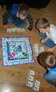 Three young boys are sitting on a wooden floor playing Santa's Rooftop Scramble, a festive board game. Game cards and pieces are spread around the colourful Christmas-themed game board.
