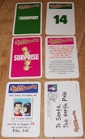 Six playing cards from the board game Santa's Rooftop Scramble are laid out on a wooden surface, featuring colourful designs with festive Christmas messages, "TRANSPORT," "14," "SURPRISE," and a postcard to Santa.