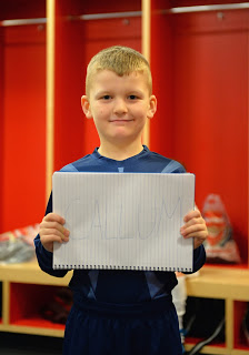 A little boy in a blue sports kit stands in a locker room, smiling and holding a notebook with "CALLUM" written on it—a dream come true. Red lockers and sports bags are visible in the background.