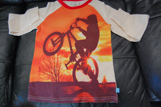 A stylish boys' shirt from Mothercare featuring a person performing a wheelie on a bicycle at sunset, set against leafless trees and an orange sky. Cream sleeves and a red collar add to its trendy look.