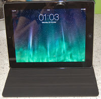 A black tablet in a Case Logic iPad case stands upright on a speckled worktop. The screen displays the time "01:03" and the date "Saturday, 26 October" over a green and blue aurora wallpaper.