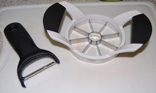 A black-handled vegetable peeler and a white apple slicer with metal blades sit on a white chopping board, ready to help you prep fruit for your favourite Apple Crumble Recipe.