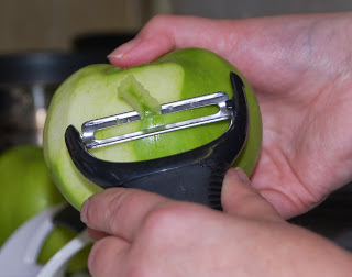 A close-up of hands holding a green apple and peeling its skin with a black vegetable peeler, perfect for starting an easy dessert like an Apple Crumble Recipe.