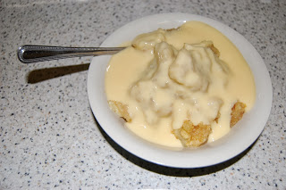 A white bowl filled with bread pudding covered in creamy custard, with a metal spoon resting inside the bowl, evokes memories of a classic Apple Crumble Recipe, all set on a speckled worktop.