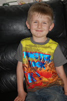 A young boy with short blond hair sits on a black leather sofa, smiling. He is wearing a colourful t-shirt from the latest Stylish Boys' Clothes collection, featuring bold, graphic text and illustrations.
