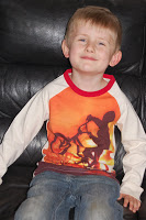 A young boy with short light brown hair smiles whilst sitting on a black leather sofa. He is wearing jeans and a stylish long-sleeved shirt from Mothercare, featuring a silhouette of a person riding a bicycle against an orange background.