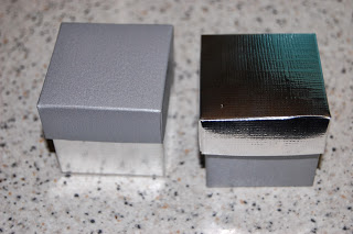 Two small square wedding favour boxes on a speckled countertop; the left box has a matte grey lid and reflective sides, while the right features a shiny silver lid and matte grey sides—perfect for paper themes.