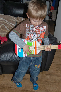 A young boy wearing a brown shirt and jeans stands indoors, looking down as he plays sweet music on a colourful ELC toy guitar. He is in front of a dark sofa on a wooden floor.