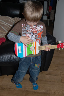A young child with blonde hair wearing glasses, a printed brown shirt, jeans, and blue socks plays a colourful ELC toy guitar, making sweet music whilst standing on a wooden floor next to a black sofa.