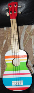 A small ELC ukulele with a red headstock and body decorated with colourful horizontal stripes in white, orange, green, blue, and pink rests on a patterned cushion—ready to create sweet music.