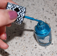 A hand with painted nails holds a brush, applying blue glittery nail varnish from an open bottle on a speckled worktop, perfect for those who love pampering or are searching for unique Wilko gifts.