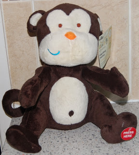 A brown and white stuffed monkey toy from Wilko Gifts sits on a kitchen worktop. The toy has a tag on its ear and a red circle on its foot that says "PRESS HERE." Beige tiled walls can be seen in the background.