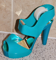 A teal Scotch Magic Tape Dispenser, shaped like a high-heeled shoe, sits on a speckled worktop with a beige tiled background.