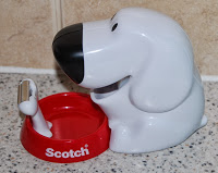 A Scotch Magic Tape Dispenser shaped like a white dog with a black nose sits next to a red bowl labelled "Scotch" on a speckled worktop.