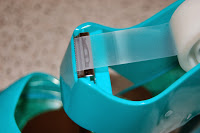 Close-up of a blue Scotch Magic Tape Dispenser holding a roll of clear adhesive tape, with the tape pulled out and ready to be cut on the metal serrated edge. A blurred surface is visible in the background.