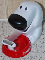 A Scotch Magic Tape Dispenser shaped like a white dog with a black nose and eyes holds a roll of clear tape in its red base labelled "Scotch," sitting on a speckled worktop.