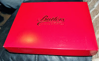 A red rectangular box from the Butlers Christmas Chocolate Collection, with "Butlers Chocolates" in gold cursive on the lid, rests on a dark leather surface.