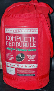 A red pack labelled "Complete Bed Bundle" with white and green text, featuring John Cotton quality and holiday designs. The cylindrical bundle with a handle includes bedding essentials, making it ideal for Christmas guests or as a White Christmas Duvet Set.