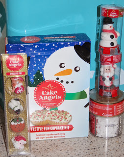 A festive Cake Angels cupcake kit features a snowman design box, a pack of decorated Christmas cupcakes, and two clear tubes with snowman and Father Christmas cake toppers, all beautifully arranged on a blue background.