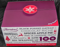 A purple and white Thorntons Christmas box featuring festive phrases like "Black Forest Gateau," "Spiced Apple Pie," and "Christmas Indulgence," topped with a red snowflake gift tag.