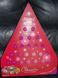 A red, triangular Thornton’s Christmas Classics Advent Calendar decorated with numbered baubles and small stars, featuring assorted chocolates pictured at the bottom. The calendar is placed on a dark background.