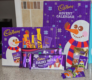 Assorted Cadbury Christmas treats and chocolates are displayed in front of two purple Cadbury Advent calendars decorated with snowmen and snowflakes, set on a kitchen worktop.