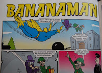 Comic panel titled "BANANAMAN" from the 2014 Beano. A superhero in blue and yellow flies overhead as two villains in purple, one holding a green bag, stand by a building labelled "Bank." A police officer points and speaks—perfect gift idea!.