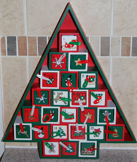 A triangular, Christmas tree-shaped advent calendar with 24 small, numbered drawers, decorated with festive holiday images and classic Country Basket Christmas decorations. The calendar stands against a tiled wall.