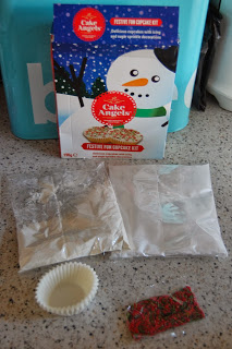 A festive Cake Angels cupcake kit with a snowman box, cupcake cases, cake mix, white icing mix, and red and green sprinkles adds holiday cheer to your kitchen worktop.
