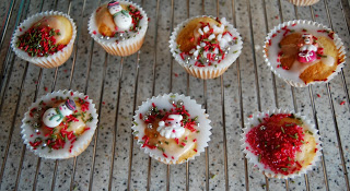 Seven decorated cupcakes sit on a cooling rack, each topped with white icing, red and green sprinkles, and festive cake angels alongside snowmen and Father Christmas figures for a charming holiday display.