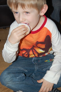 A young boy in a red and white shirt with a bicycle graphic sits on the floor, holding and eating a fairy cake with a white wrapper, savouring every bite like one of the little cake angels.