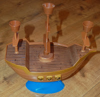 A plastic toy pirate ship from Don't Rock the Boat, with a brown deck and three upright funnels, sits on a blue base placed on a wooden floor.