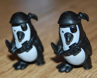Two small toy penguins dressed as pirates, each wearing a black helmet, black eye patch, and a belt, stand on a wooden surface—ready for adventure in your next game of Don't Rock the Boat.