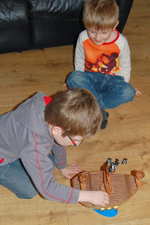 Two young boys play on a wooden floor with a toy pirate ship, careful not to tip it like in Don't Rock The Boat. One boy, wearing glasses and a grey hoodie, moves pieces while the other, in a colourful shirt, sits cross-legged and watches closely.
