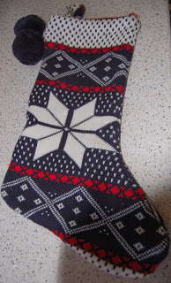 A knitted Christmas stocking with a white snowflake pattern, red accents, and two pom-poms, from HomeSense, lying on a speckled white surface.