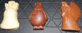 Three pieces of moulded chocolate from Hotel Chocolat’s Mingles and Jingles collection rest on a cooling rack: a white chocolate bell, a milk chocolate penguin in a scarf, and a turkey with fanned tail feathers—perfect for any festive chocolate bag.