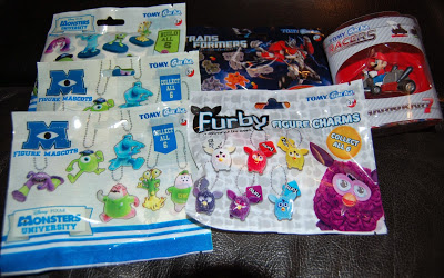 A variety of toy blind bags, including Gacha-style Monsters University, Furby figure charms, Transformers, and Cars characters, are displayed on a dark surface. The packaging reveals the collectable items inside each series.