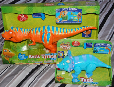 Two toy dinosaur figures in colourful packaging: an orange and blue Tyrannosaurus named Boris and a blue Triceratops named Tank, both from the show "Dinosaur Train," make perfect Christmas presents, displayed on a striped surface.