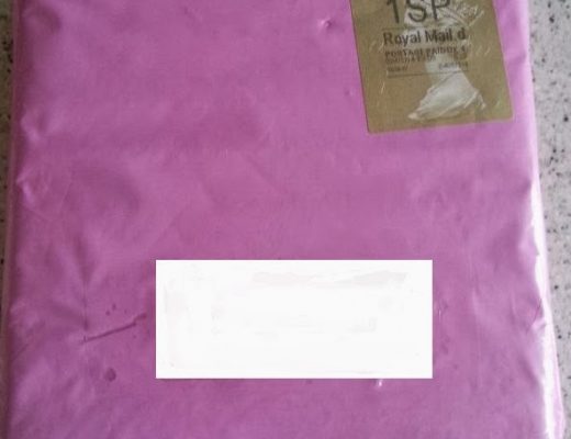 A square parcel wrapped in pink paper with a gold Royal Mail "1st" sticker sits on a speckled surface—perfect as a unique Valentines present. The address label in the centre is blanked out.
