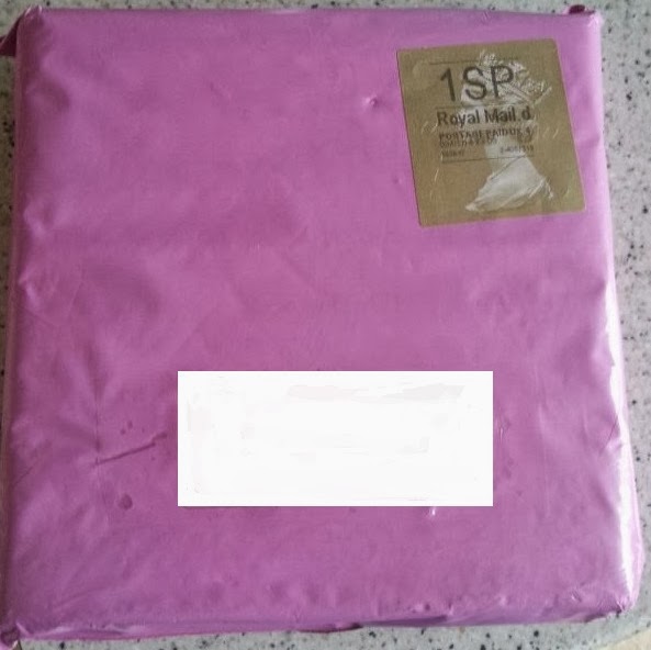A square parcel wrapped in pink paper with a gold Royal Mail "1st" sticker sits on a speckled surface—perfect as a unique Valentines present. The address label in the centre is blanked out.