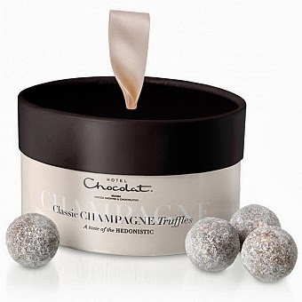 A round, light-coloured box of Hotel Chocolat Classic Champagne Truffles with a dark lid and ribbon handle is shown. Perfect for a Valentines Giveaway, four powder-dusted chocolate truffles are placed outside the box.