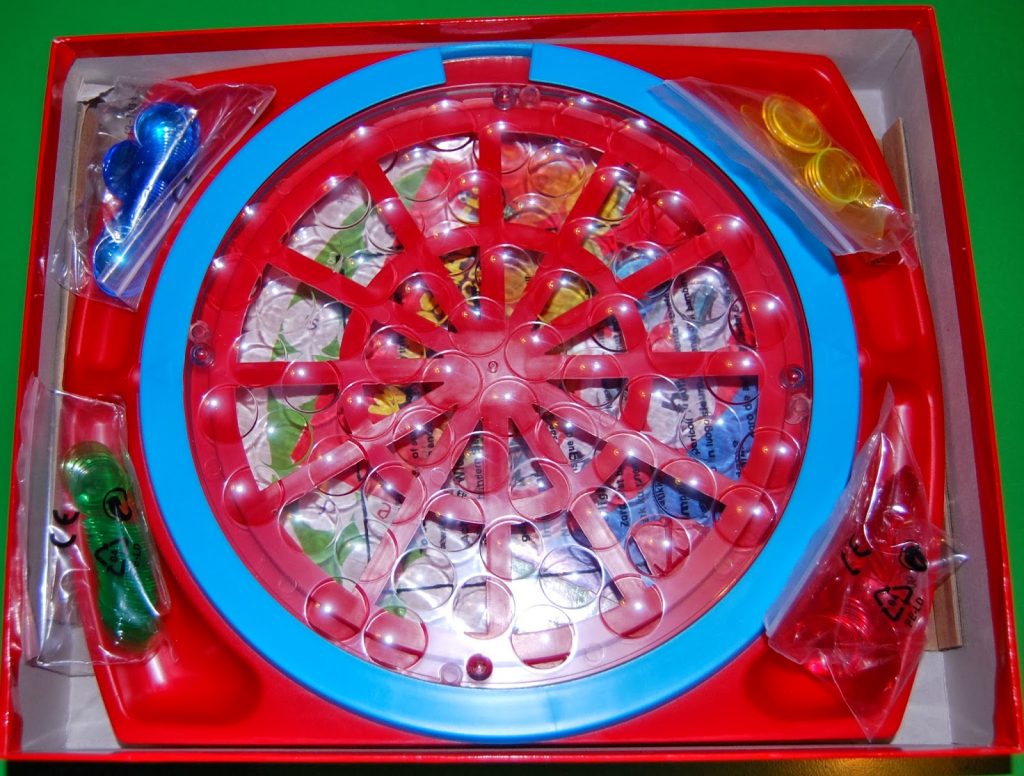 A board game featuring a red and blue circular plastic frame with multiple slots, four bags of colourful game pieces, and a printed Wordsearch Junior game board underneath, all inside an open box.