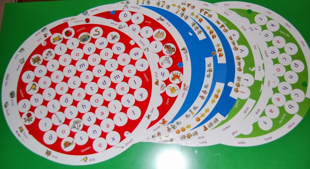 Colourful circular learning cards with alphabet letters and small pictures of objects and animals, inspired by Wordsearch Junior, are arranged on a green surface. The cards are partially stacked and fanned out, perfect for fun competition or educational review.